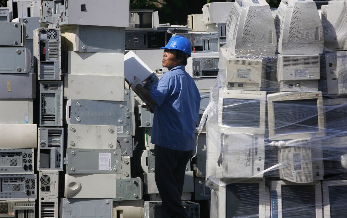 Worker in blue hard hat and jacket inspecting stacked computer equipment and electronic waste.