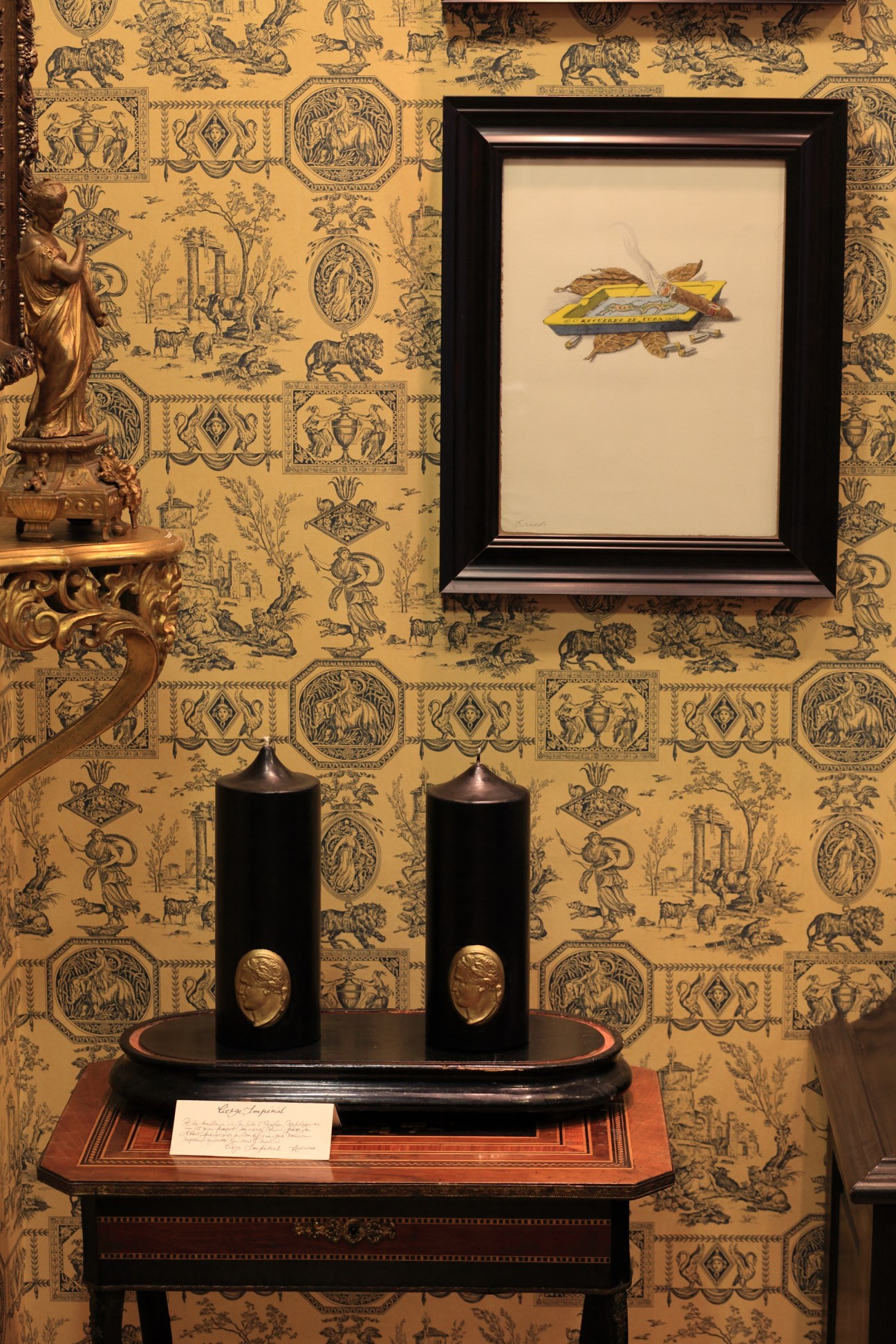 Two black pillar candles with bronze medallion profiles displayed on an ornate wooden table against patterned wallpaper.