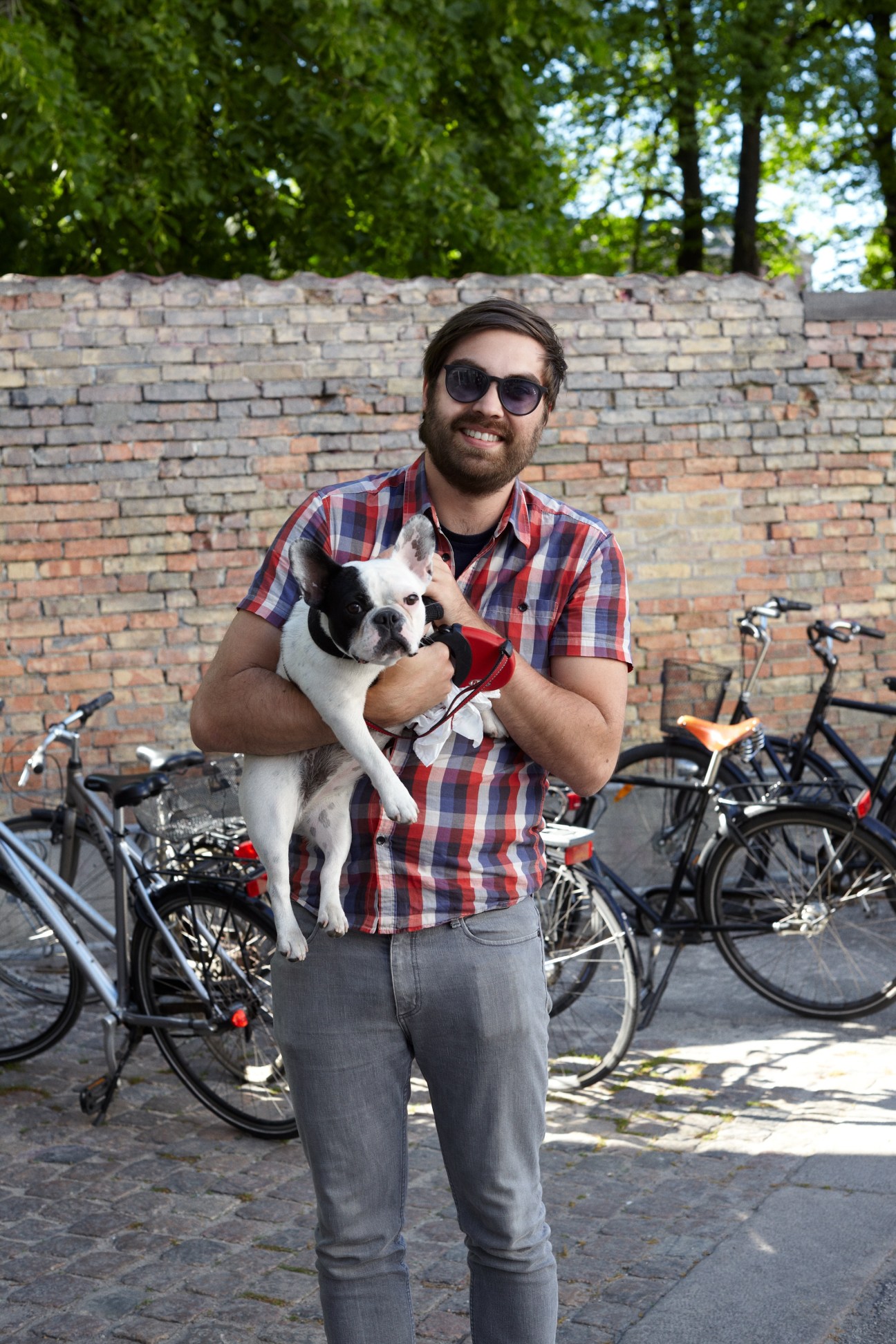 Man holding a small black and white dog against a brick wall with bicycles in the background.