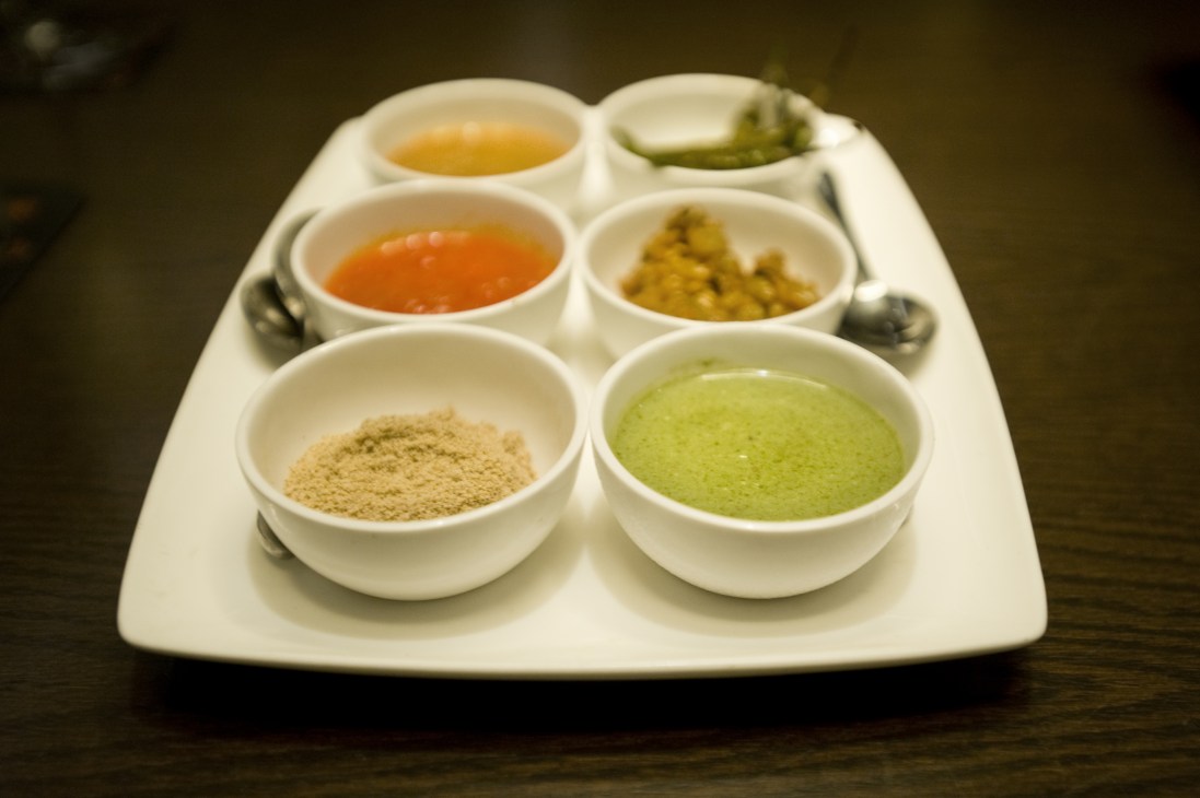 Six small white bowls containing various spices and condiments in powdered, paste, and liquid forms arranged on a white plate.