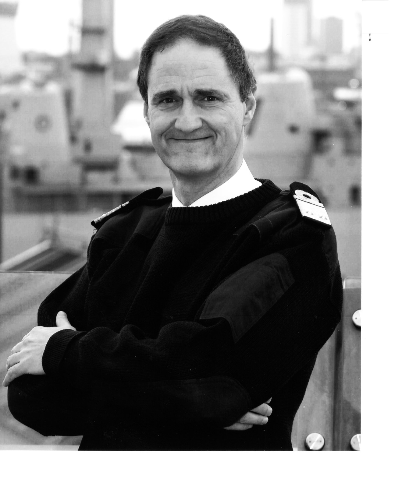 Rear Admiral Hank Ort in naval uniform standing with arms crossed before a naval vessel backdrop.