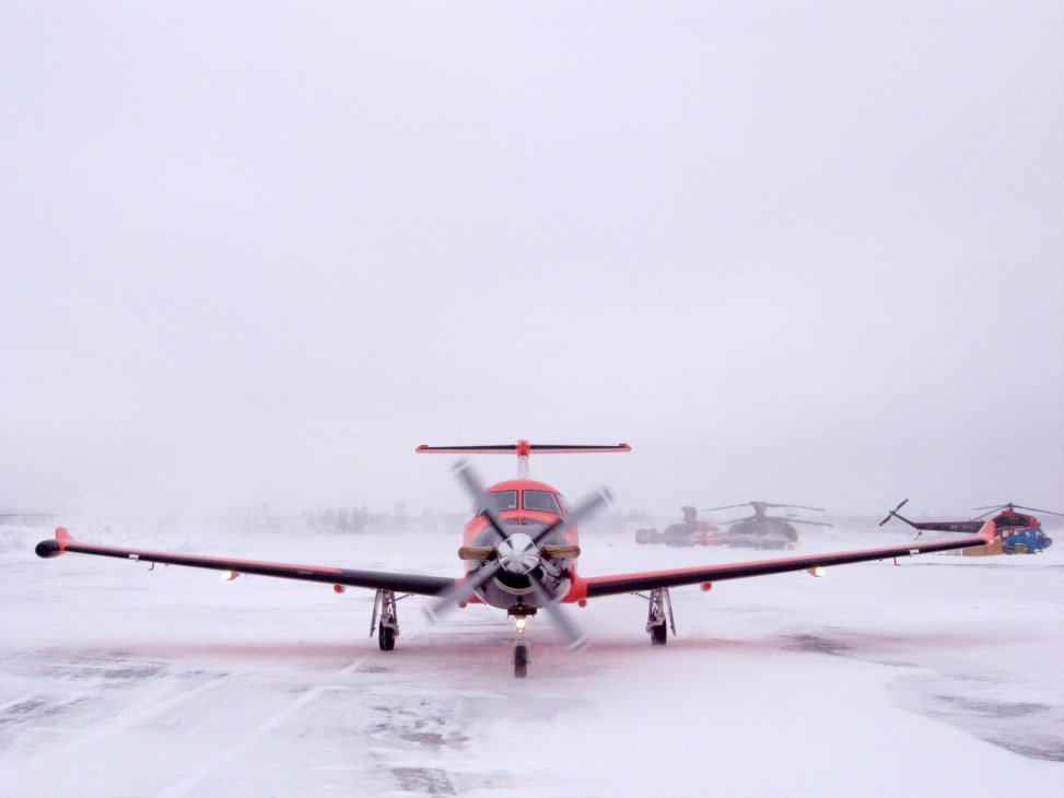Bright orange Pilatus PC-12 turboprop on snowy runway with helicopter in background.