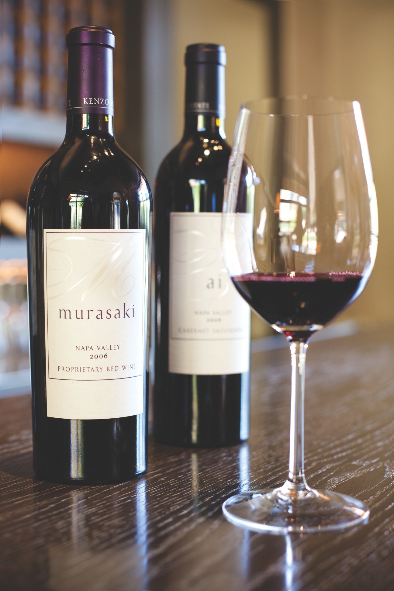Kenzo Estate wine bottles Murasaki and Ai displayed with a wine glass on a wooden table.