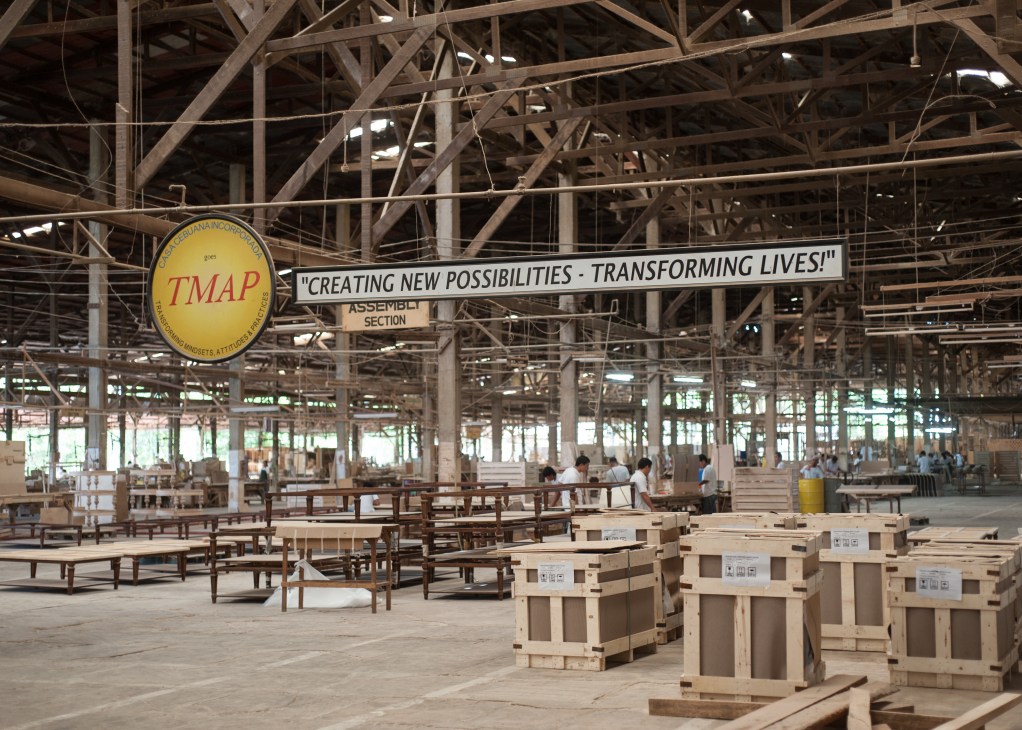 Interior of a large factory space with exposed wooden beams, stacked wooden furniture in cardboard boxes, and a yellow TMAP banner overhead.