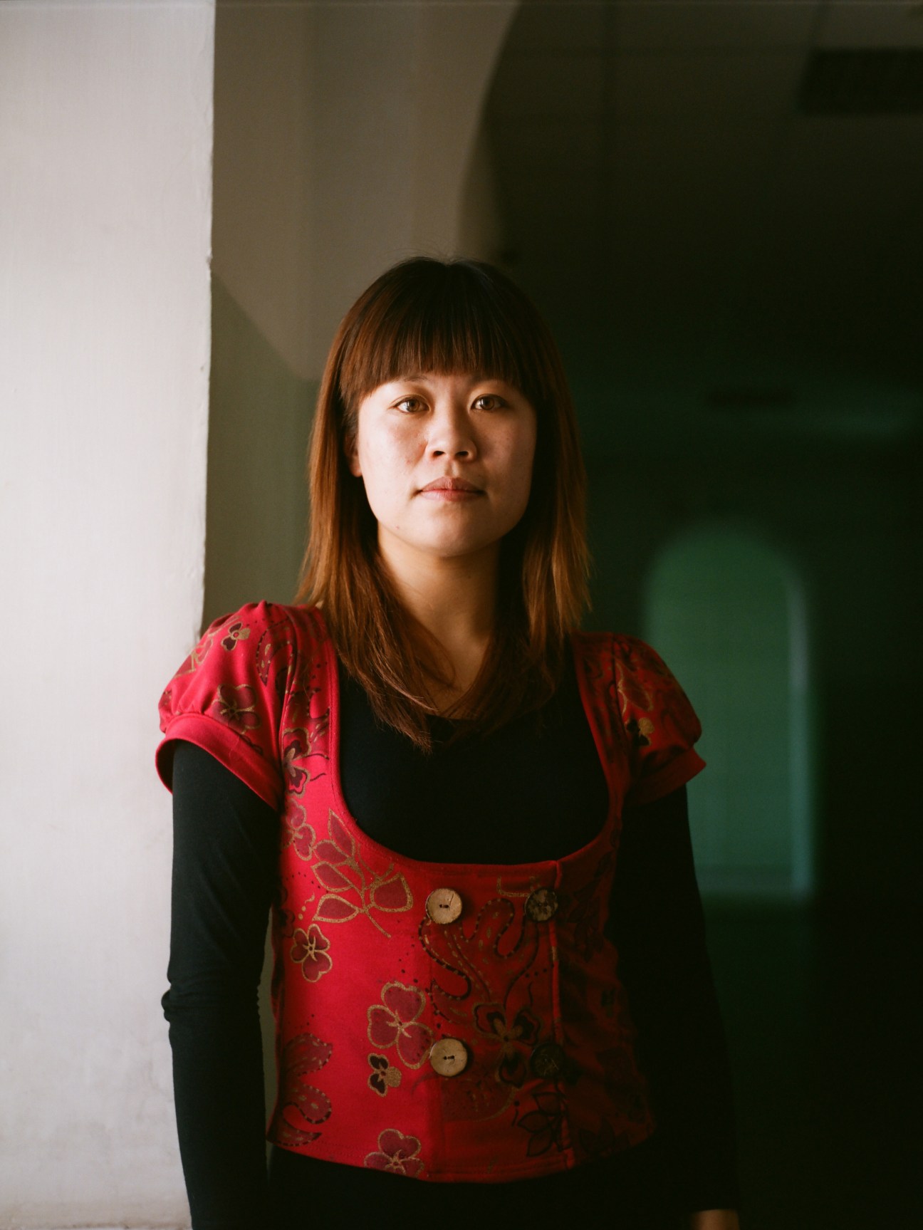 Woman wearing red patterned vest over black long-sleeved shirt standing against white and green wall.