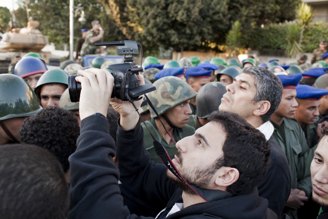 Man filming with a video camera surrounded by soldiers wearing helmets during a public gathering.