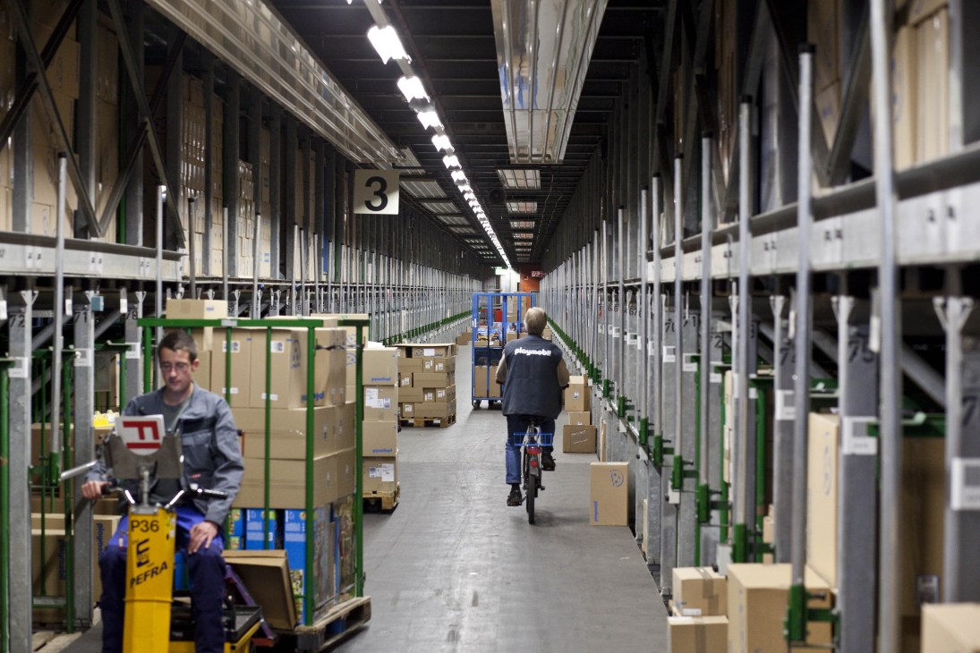 Playmobil toy factory warehouse with workers organizing boxes along metal shelving aisles.