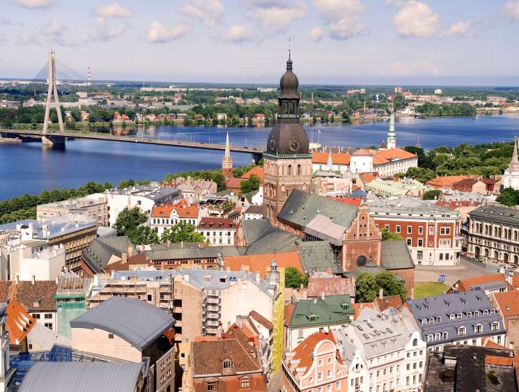 Aerial view of Riga's skyline with the Daugava River, featuring historic architecture, a cathedral dome, and modern bridges.