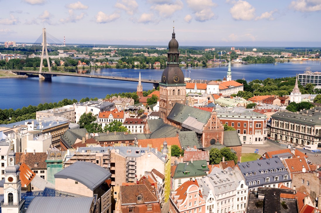 Aerial view of Riga's skyline with the Daugava River, featuring historic architecture, a cathedral dome, and modern bridges.
