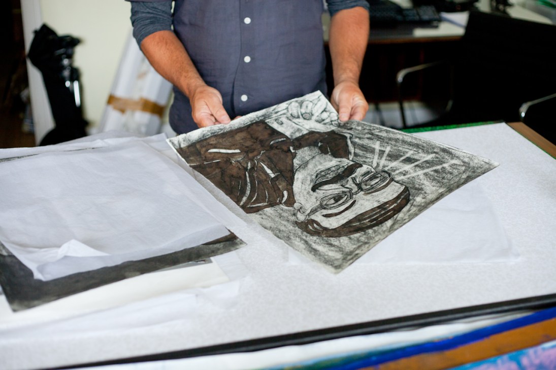 Person in blue shirt holding an etching or print featuring expressive faces and figures on white paper.