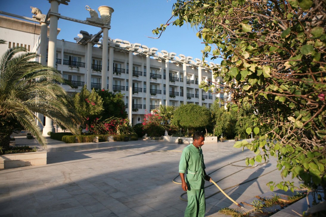 A man in green work clothes sweeps the courtyard of the Dariush hotel, a white modernist building with palm trees and flowering plants.