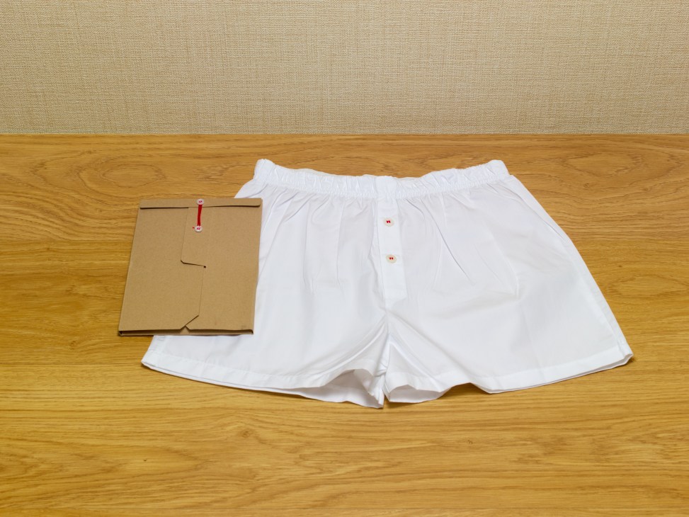 White boxer shorts with red thread on shell buttons displayed next to cardboard packaging on wooden floor.