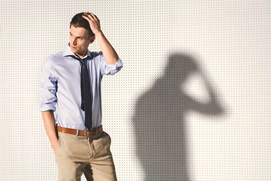 Man wearing light blue shirt with dark patterned tie and tan chinos against dotted background.