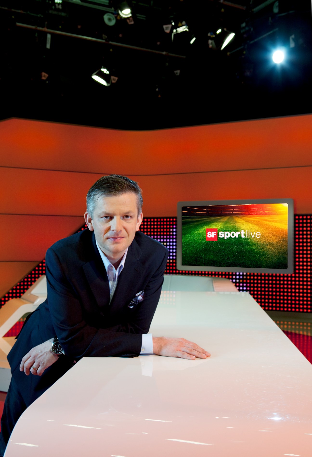 Rainer Maria Salzgeber at a TV studio desk with SF sportlive branding visible on screen behind him.