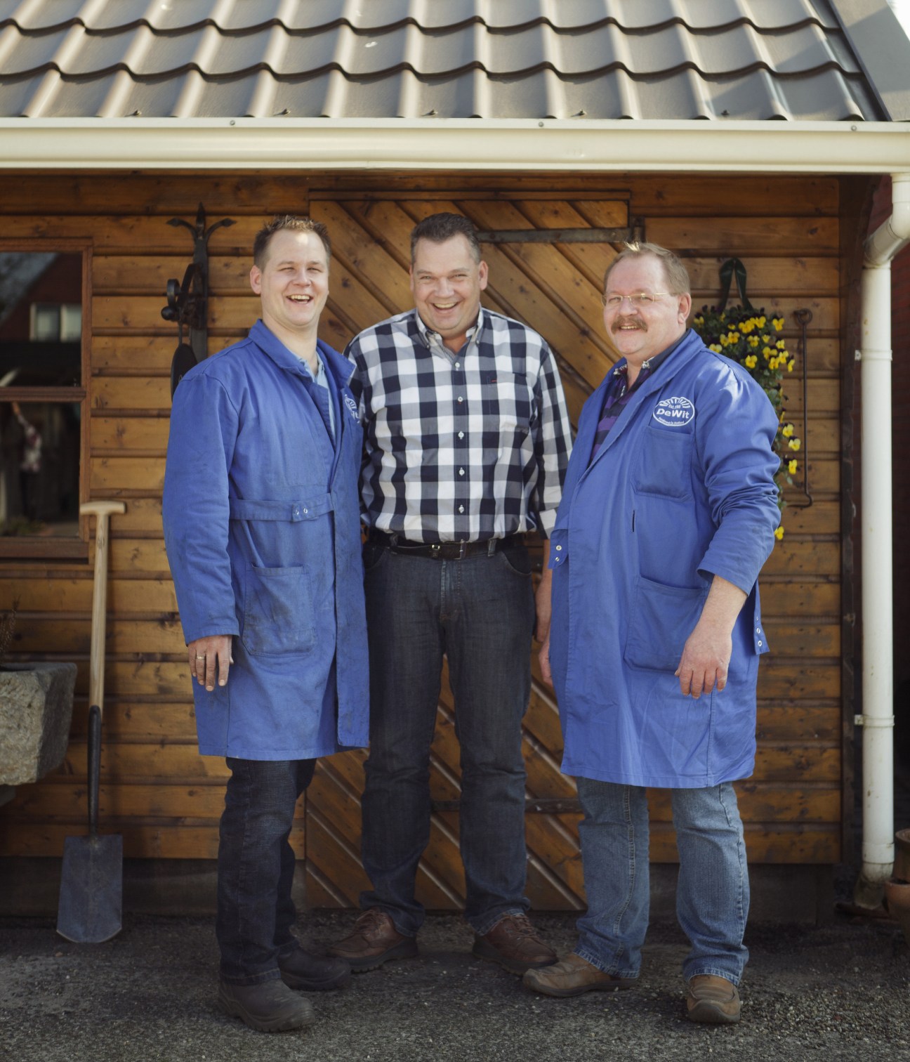Three men in blue work coats stand smiling outside a wooden building with yellow flowers.