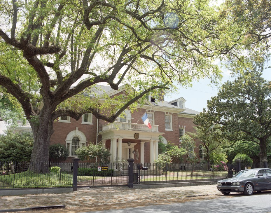 Brick mansion with white columns and French flag, shaded by large oak tree.