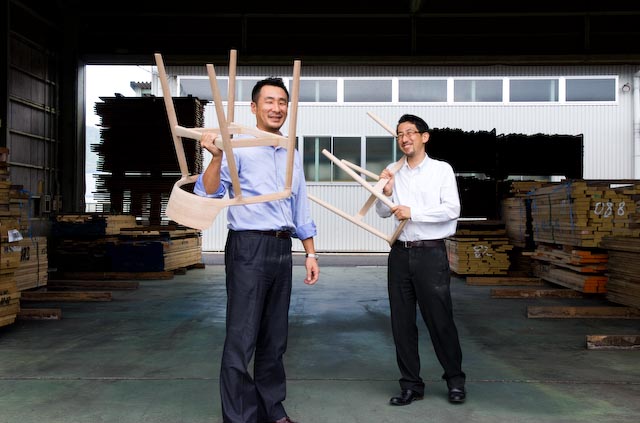 Hiroshi and Yoichi Yamanaka holding wooden chair frames in a factory yard.