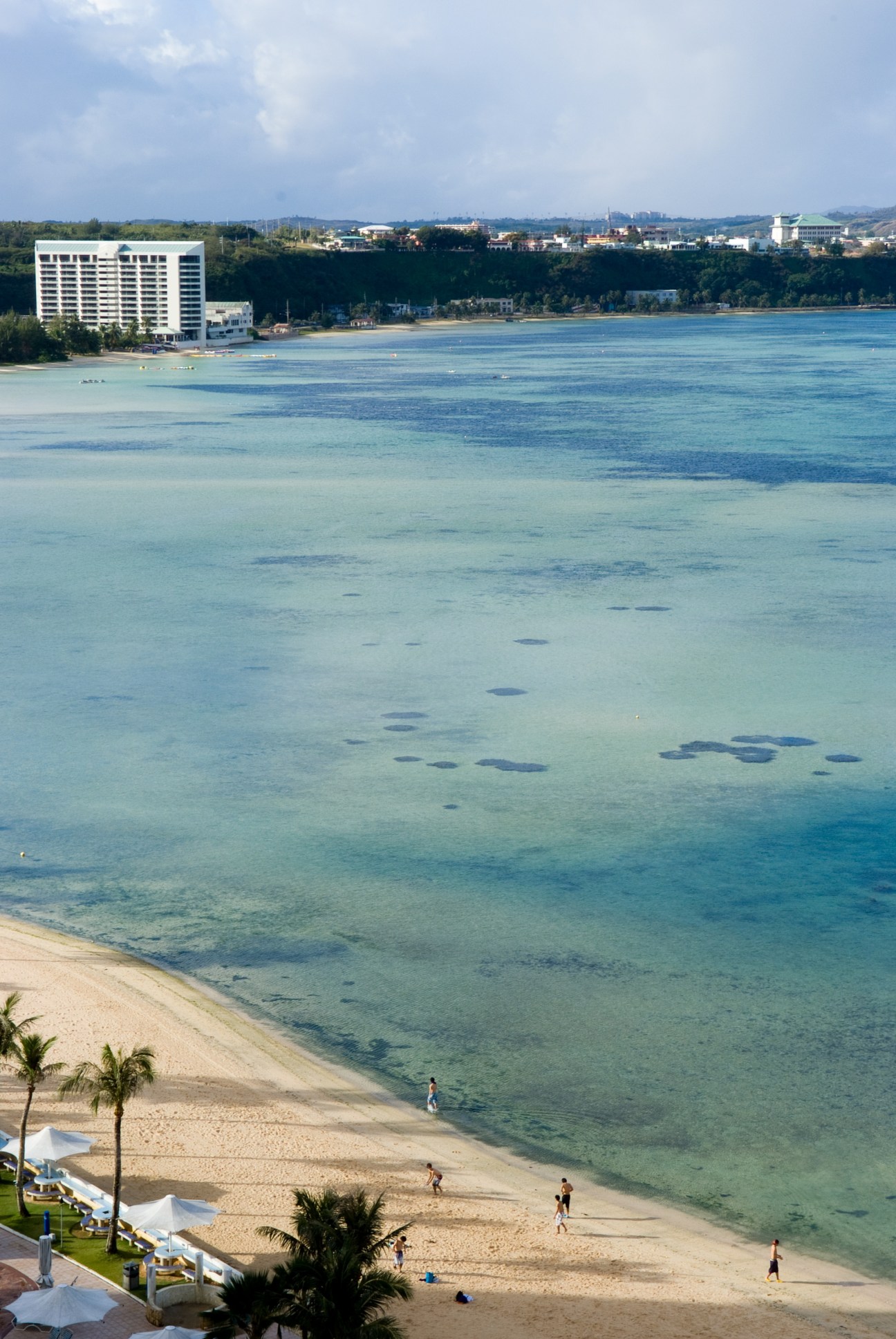 Turquoise waters and sandy beach on Guam's west coast with resort buildings and palm trees.