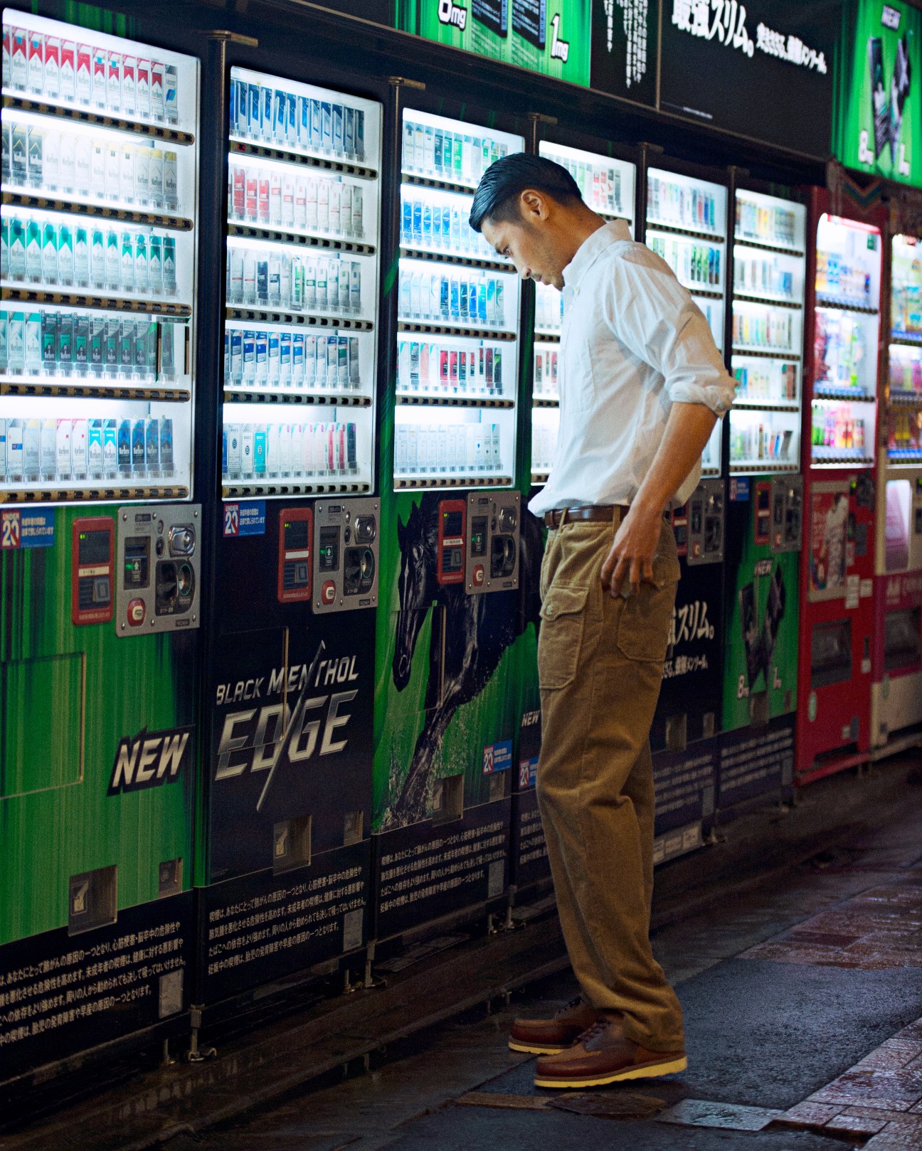 Man in white shirt and khaki trousers browsing illuminated beverage vending machines in Tokyo.