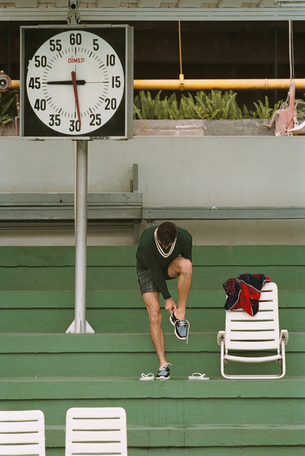 Model in green knit and patterned shorts stretches at a tennis court with a clock and lounge chair visible.