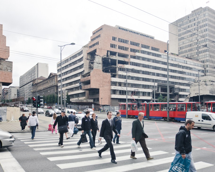 A busy downtown Belgrade street with war-damaged buildings, pedestrians at a crosswalk, and a red tram passing by.