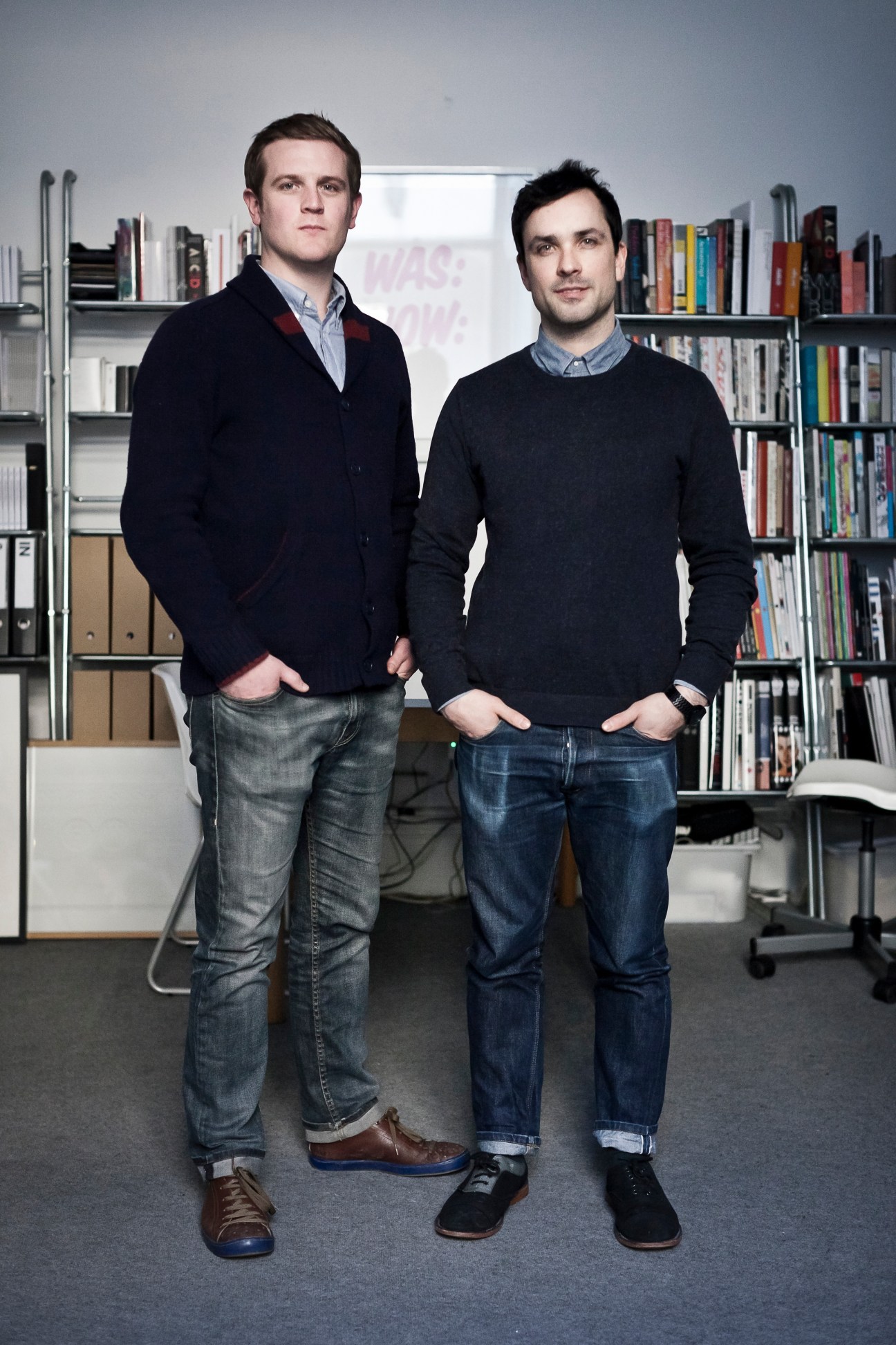 Two men standing in a design studio with bookshelves behind them.
