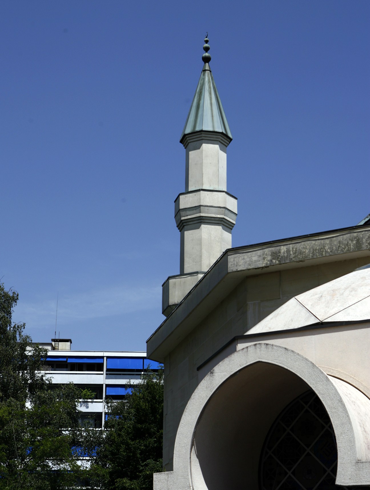 A minaret with a pointed spire rises above a modern building with blue accents against a clear blue sky.