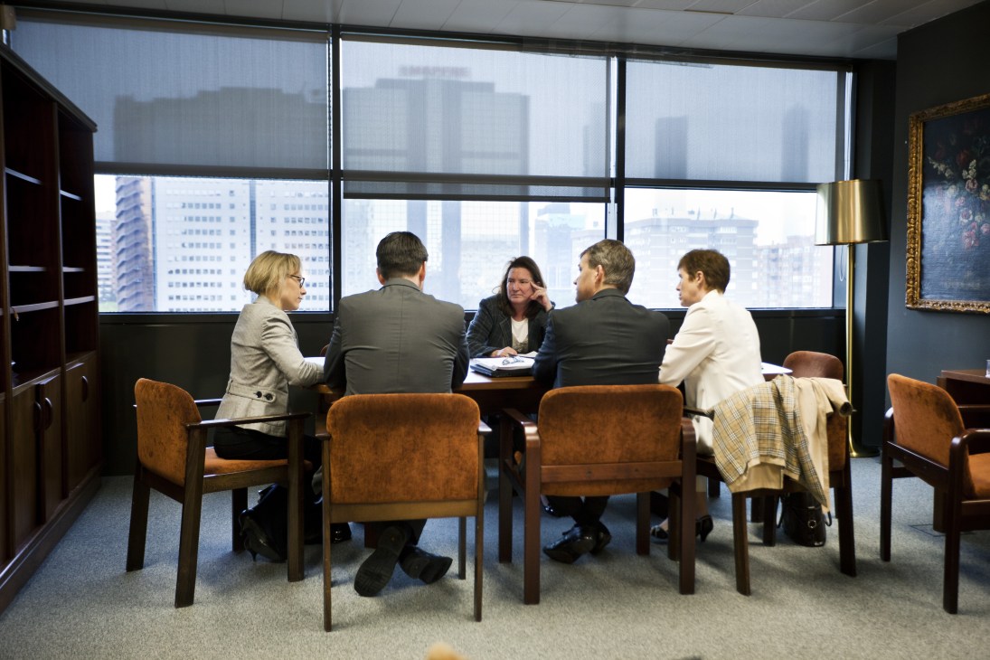 Five business professionals in formal attire sit around a table in a high-rise office with city views.