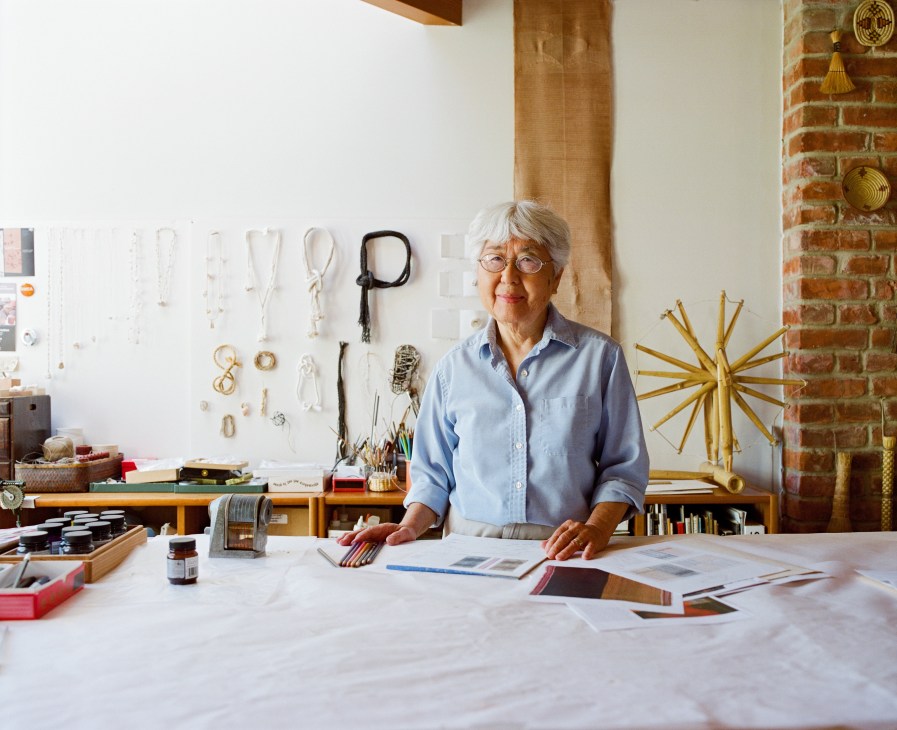 Textile artist Kay Sekimachi stands at her work table in her jewellery studio surrounded by materials and tools.