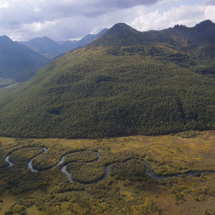 A winding river snakes through a lush forested valley with volcanic mountains in the background under cloudy skies.