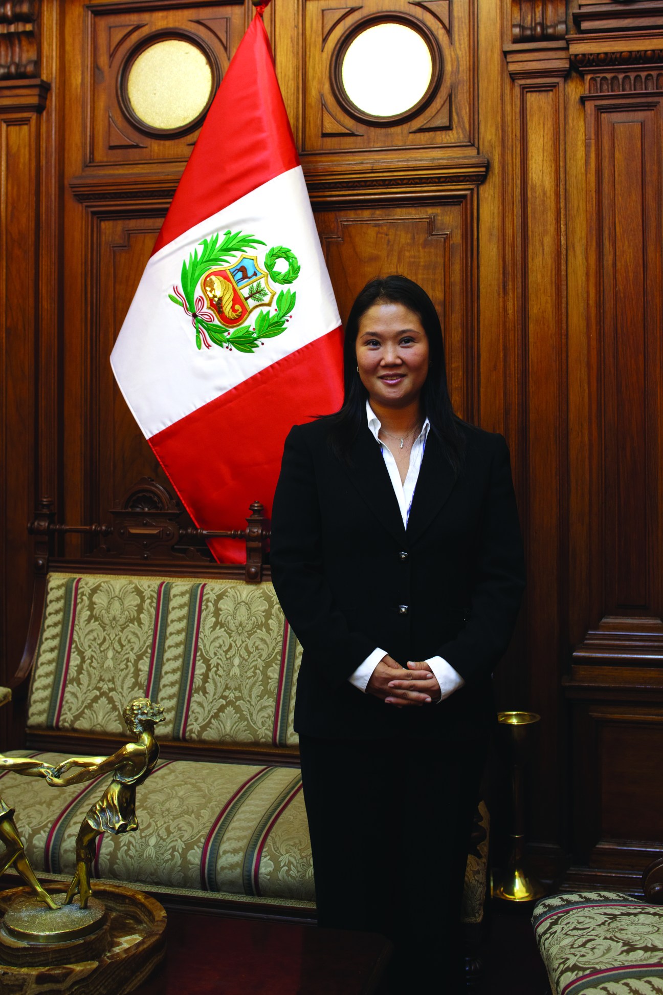 Keiko Fujimori stands in her office beside the Peruvian flag, wearing a black blazer and white shirt.