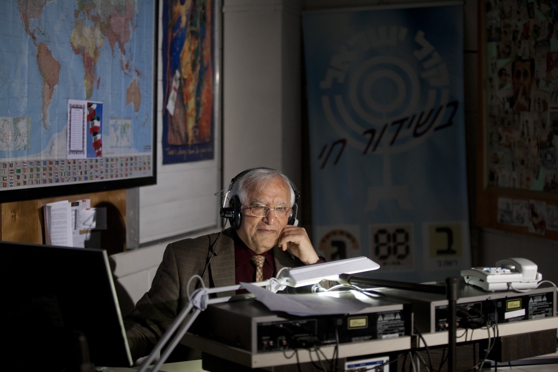 Menashe Amir wearing headphones at a radio studio desk with broadcasting equipment and a world map on the wall behind him.