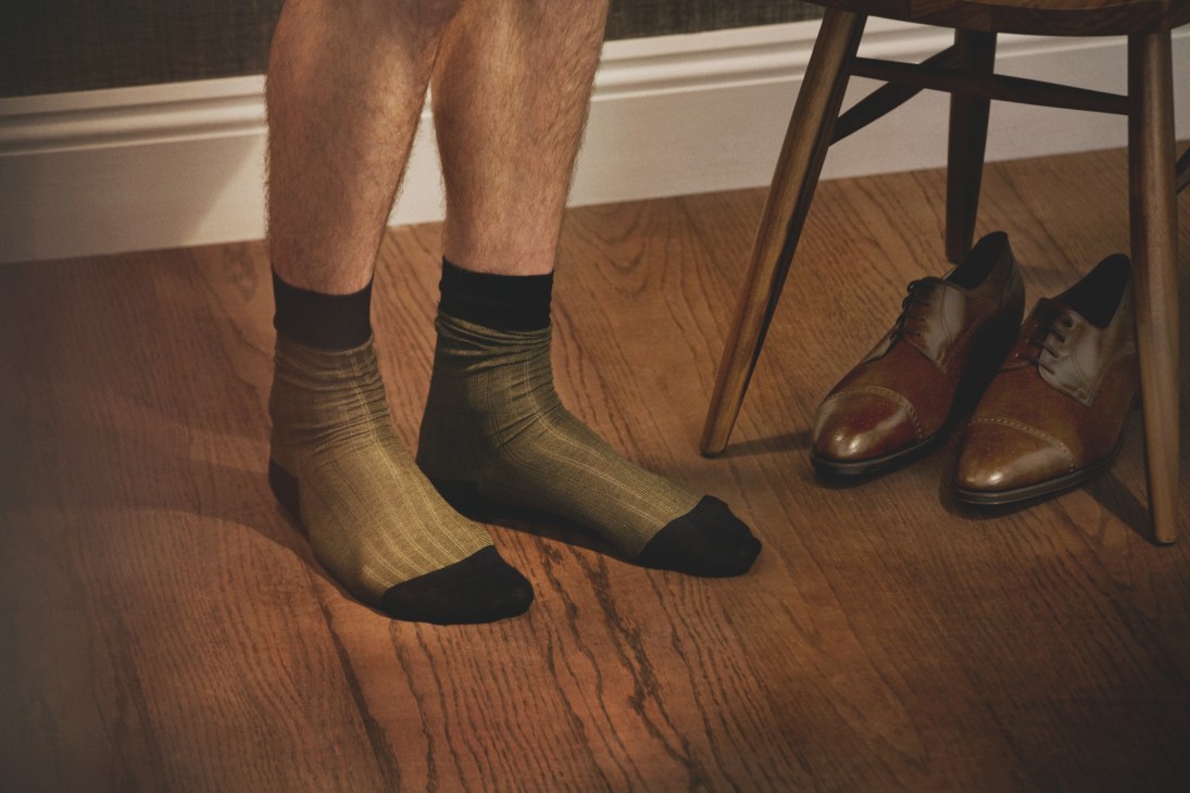 Man's legs wearing gold patterned socks by Tabio and brown leather shoes by John Lobb on wooden floor.
