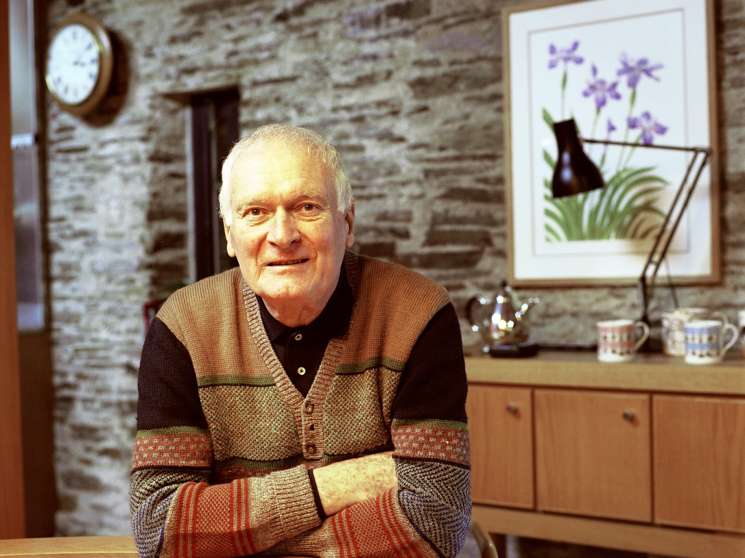 Designer Kenneth Grange seated at a wooden table in his home with exposed stone walls and modern furnishings behind him.