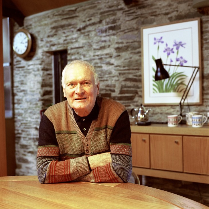Designer Kenneth Grange seated at a wooden table in his home with exposed stone walls and modern furnishings behind him.