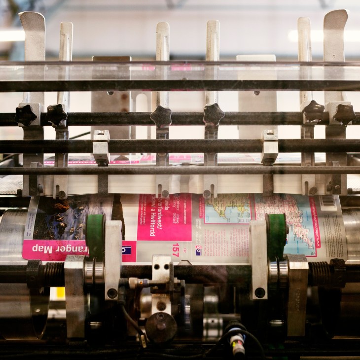 Ordnance Survey maps being printed on a lithographic press, with pink and colored map sheets visible under metal registration bars.