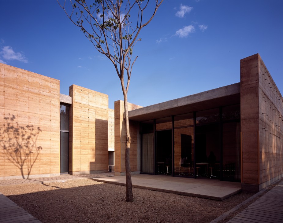 Oaxaca School of Fine Arts exterior with rammed earth walls, concrete structure, and sparse desert landscaping under blue sky.