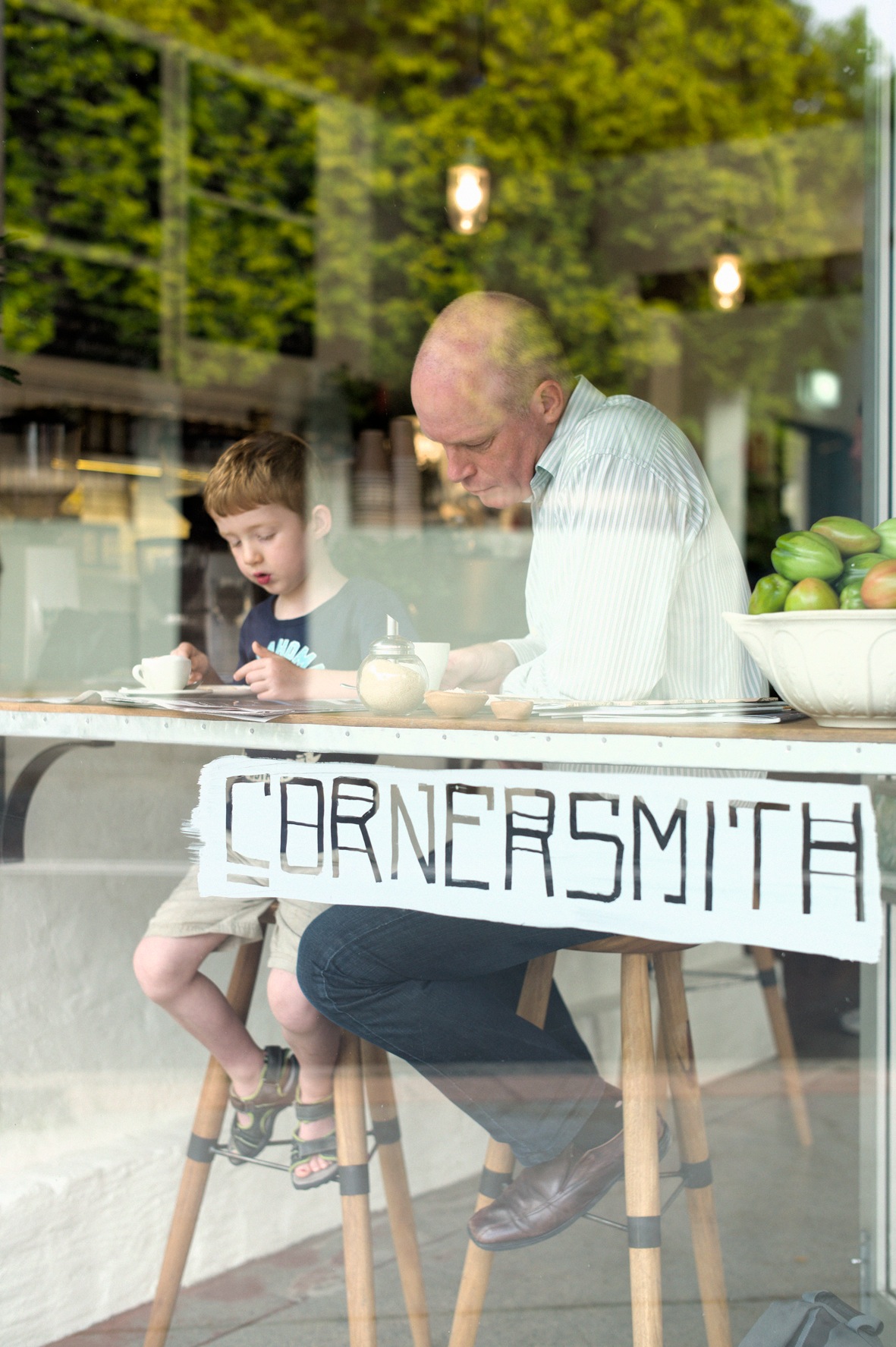 Man and child seated at Cornersmith cafe counter with fresh produce visible in background