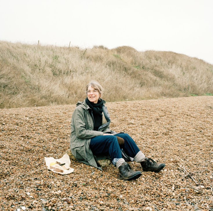 Margaret Howell sits on a shingle beach eating her picnic, wearing a green jacket and blue trousers.