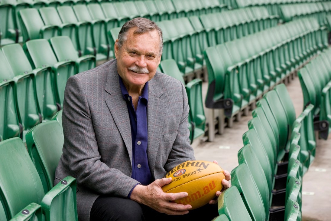 Ron Barassi holds a yellow Australian Rules football while seated in the MCG stadium stands.