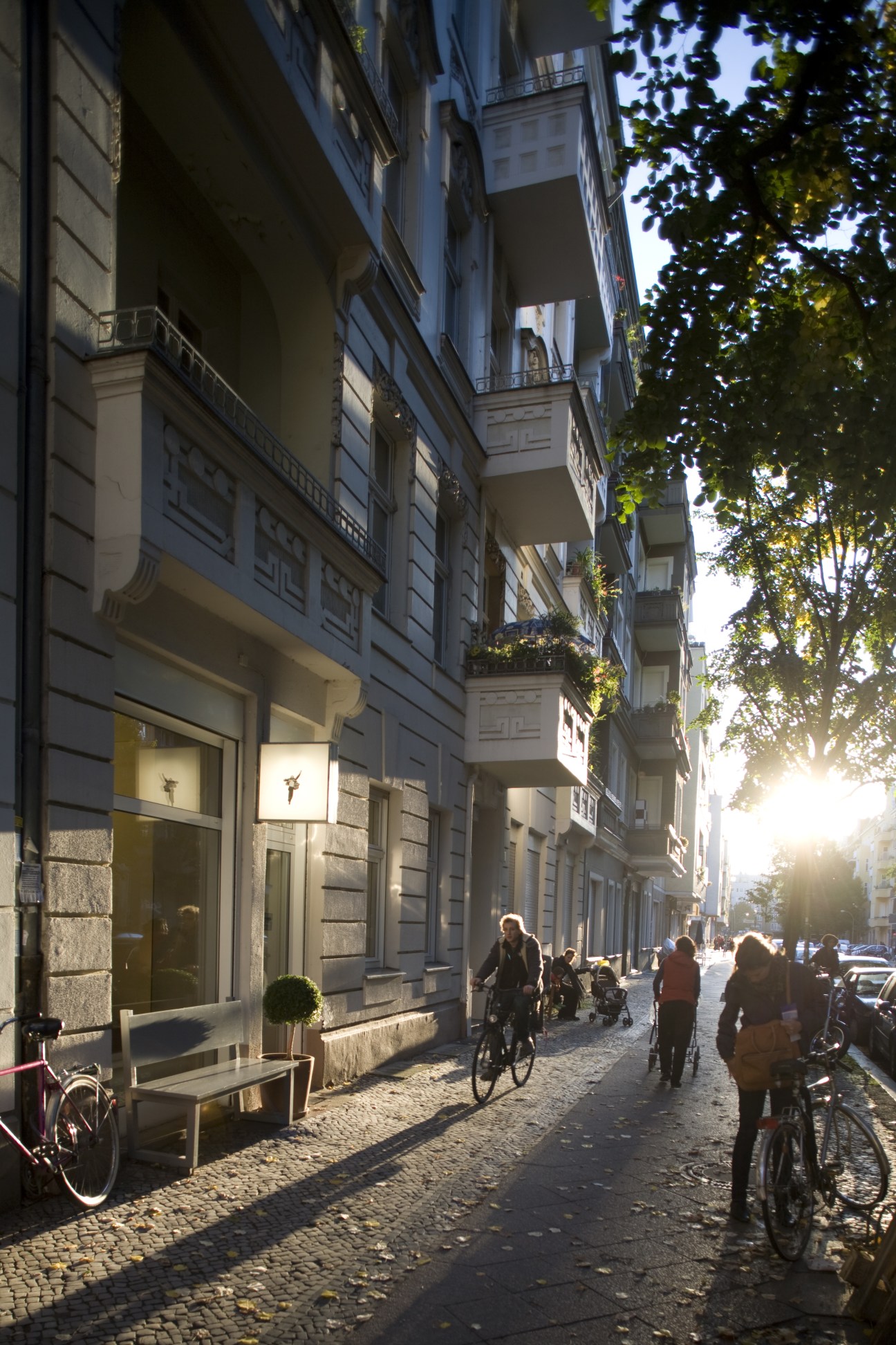 Cyclists and pedestrians on a sunny cobblestone street lined with pre-war Gründerzeit buildings and ivy-covered facades in northern Neukölln.