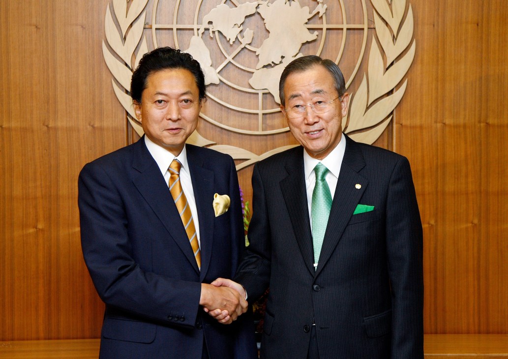 Two men in business suits shake hands in front of a wooden wall with a UN logo.