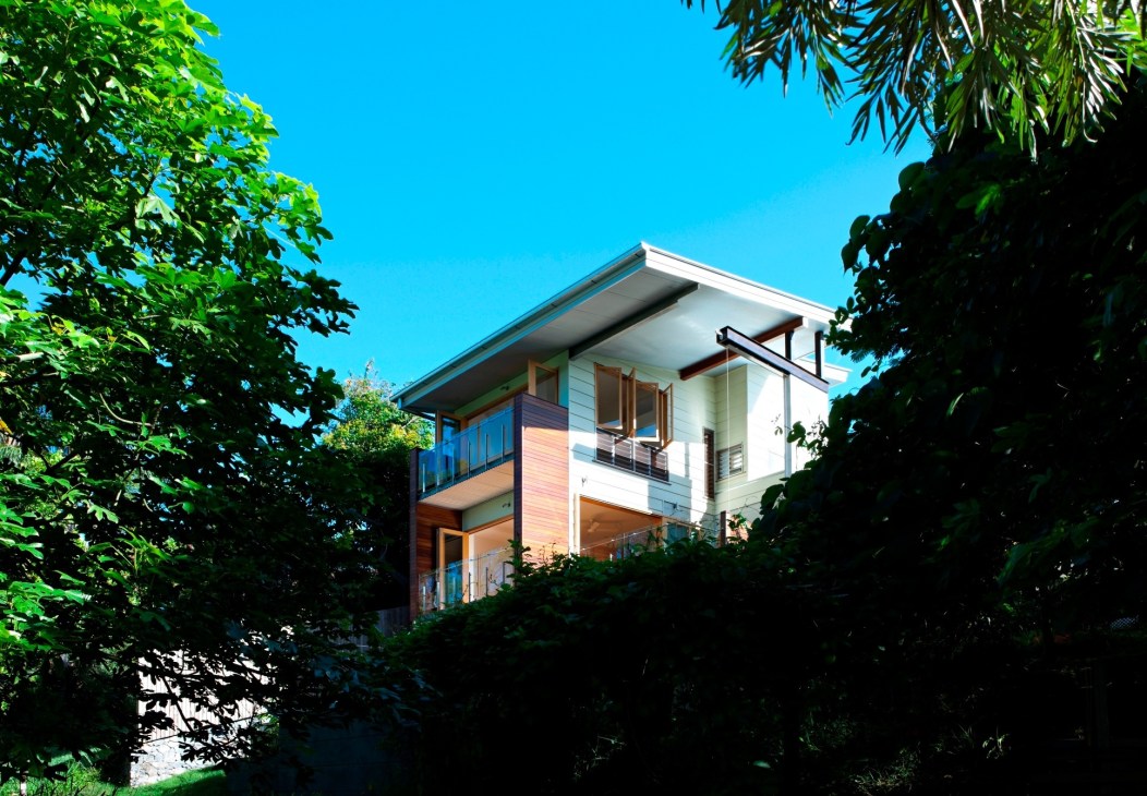 A two-story modern residential home with white and wood cladding nestled among green foliage under clear blue sky.