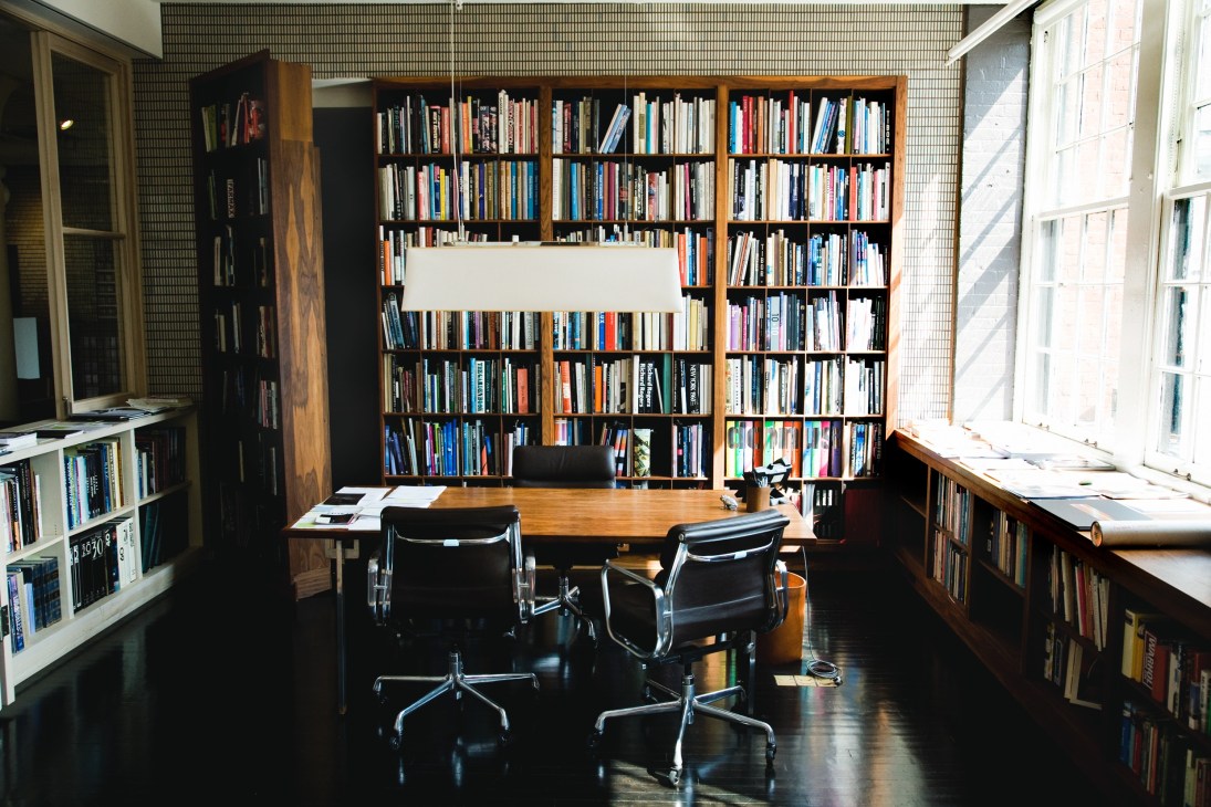 Modern office with wooden desk, black chairs, and floor-to-ceiling bookshelves filled with books and magazines.