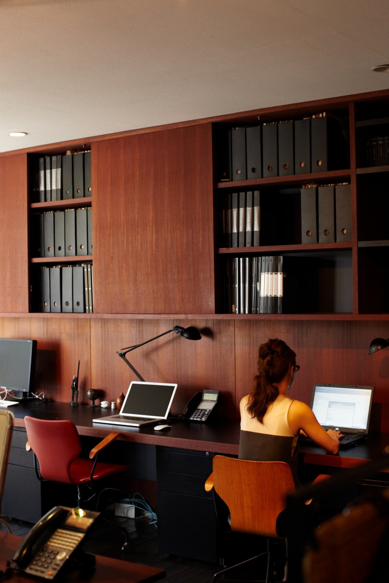 Woman working at desk in modern office with warm wood paneling and built-in shelving.