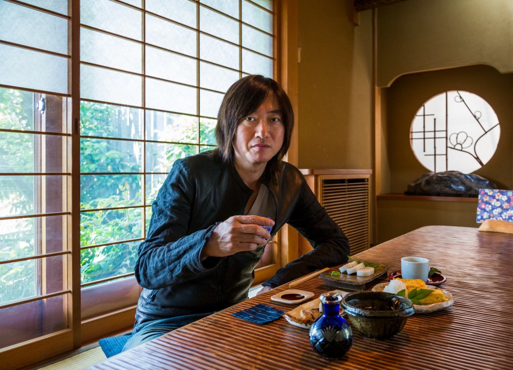 Takeshi Kobayashi sitting at a wooden counter in a traditional Japanese restaurant, holding chopsticks with food and sake on the table.