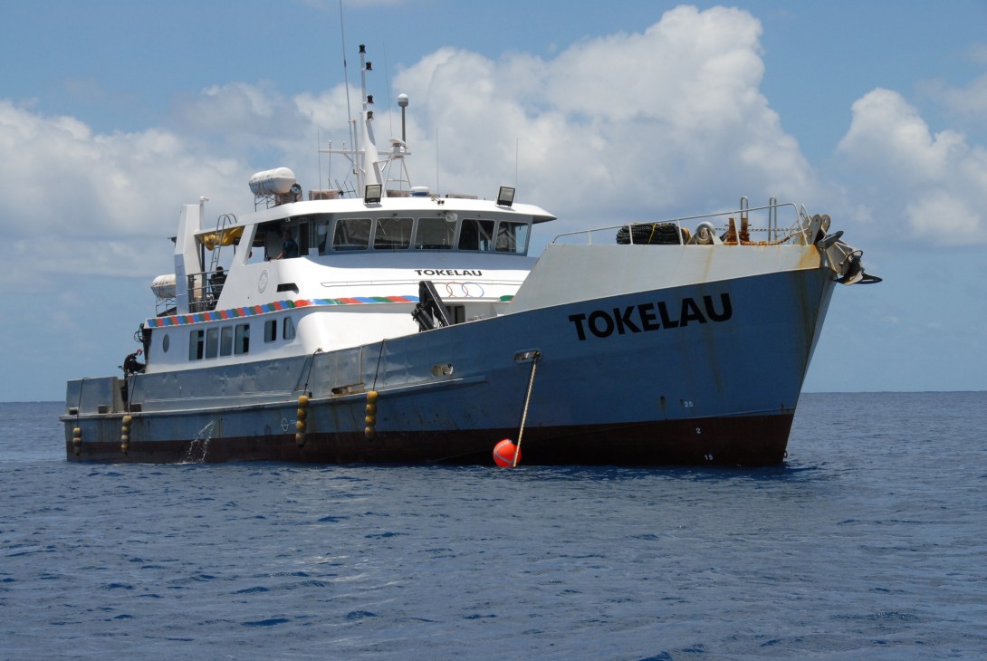 The Tokelau ferry vessel anchored in calm blue ocean waters under a cloudy sky.