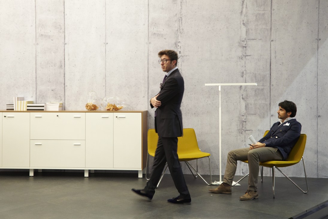 Two men in business attire at a Quadrifoglio office furniture stand with cream storage units and yellow chairs against a concrete wall.