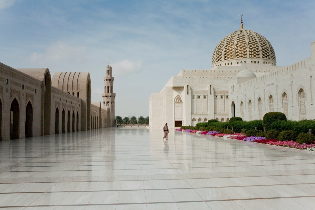 Muscat's Grand Mosque featuring white architecture with a large golden dome, courtyard with pink flowers, and a minaret.