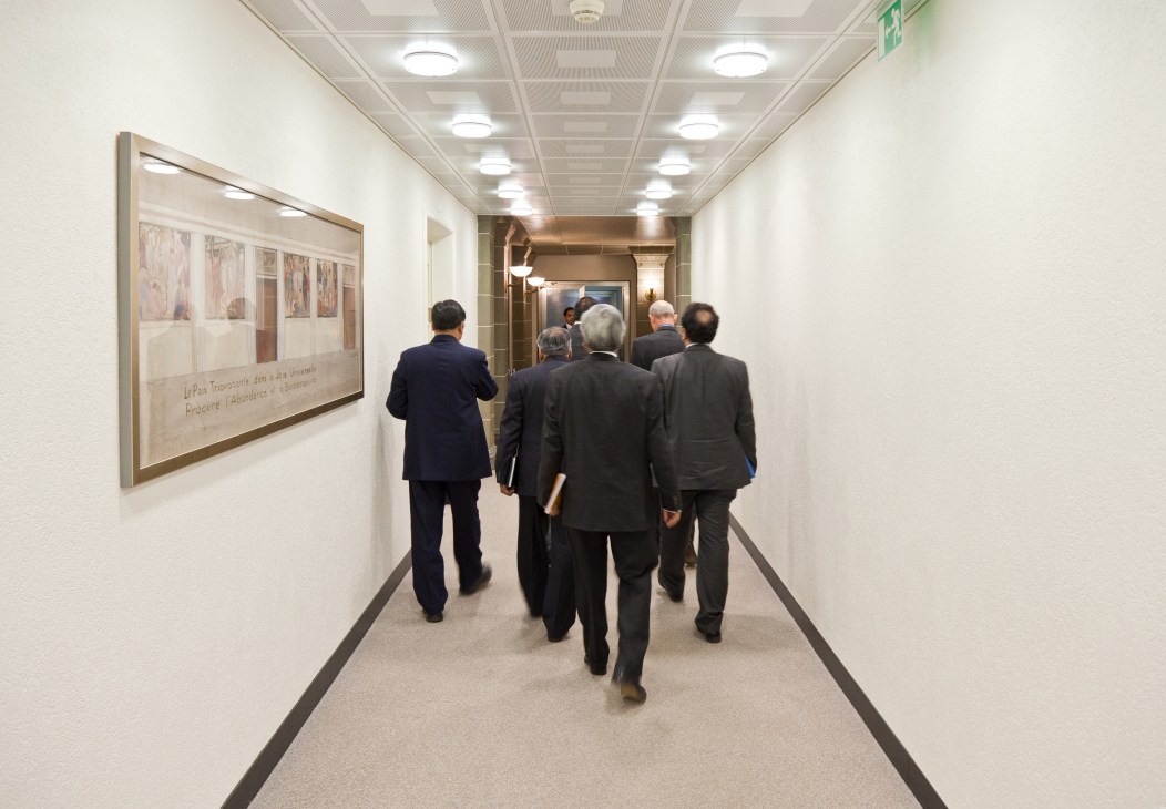Business professionals in suits walking through a modern corridor with framed artwork on the wall.